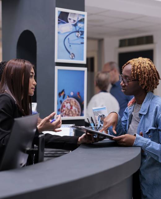 Female receptionist helping woman at reception desk, signing medical report papers to receive insurance support before checkup appointment. Patient filling in registration form to do examination.